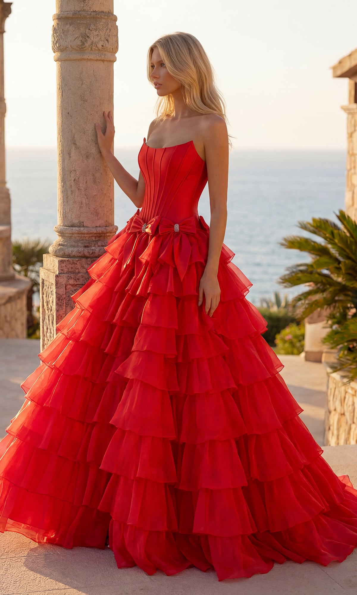 A woman stands outdoors before stone columns, wearing the PromGirl 250071 Tiered Prom Ball Gown with Bows in red. The sea and palm trees can be seen in the background.